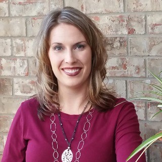 Smiling woman with light brown hair in a burgundy top standing against a brick wall.