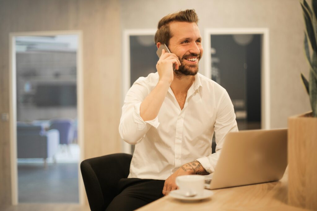 A man in a white shirt smiling while talking on the phone at a desk.