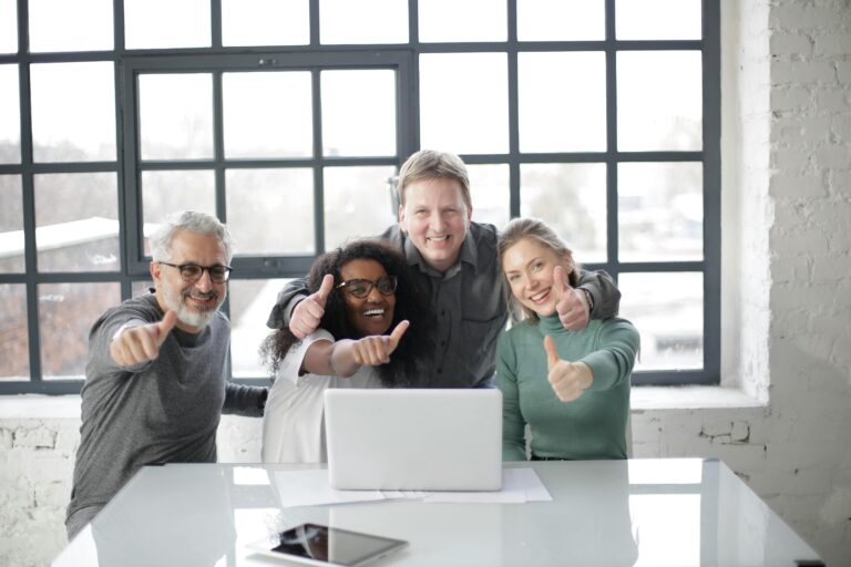 Diverse group of coworkers giving thumbs up around a laptop in a bright office.