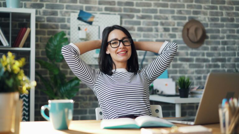 Relaxed woman with glasses smiling and leaning back at her desk.